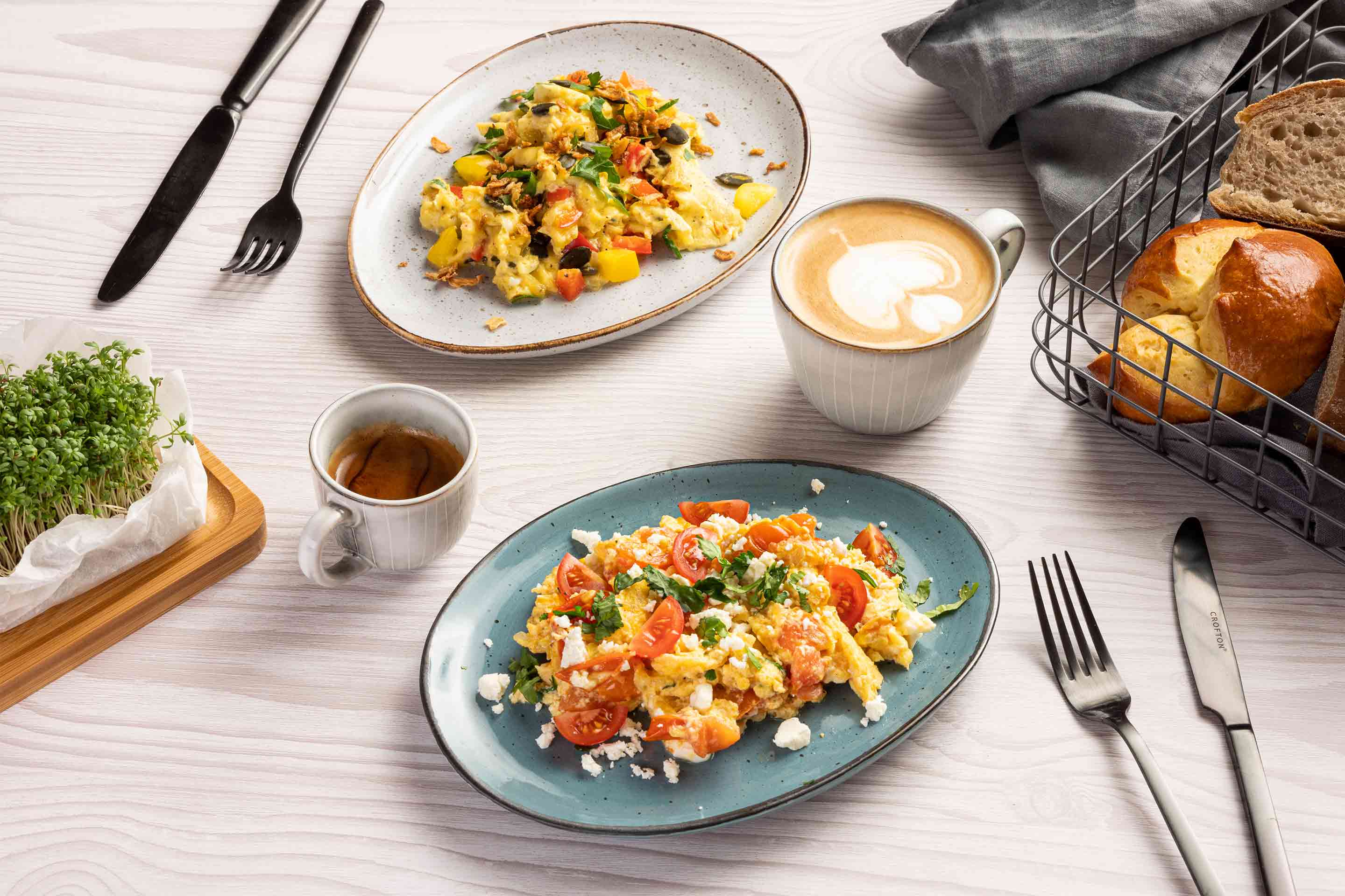 Две чинии с два вида бъркани яйца, поднесени с кафе и капучино, и кошница хляб.