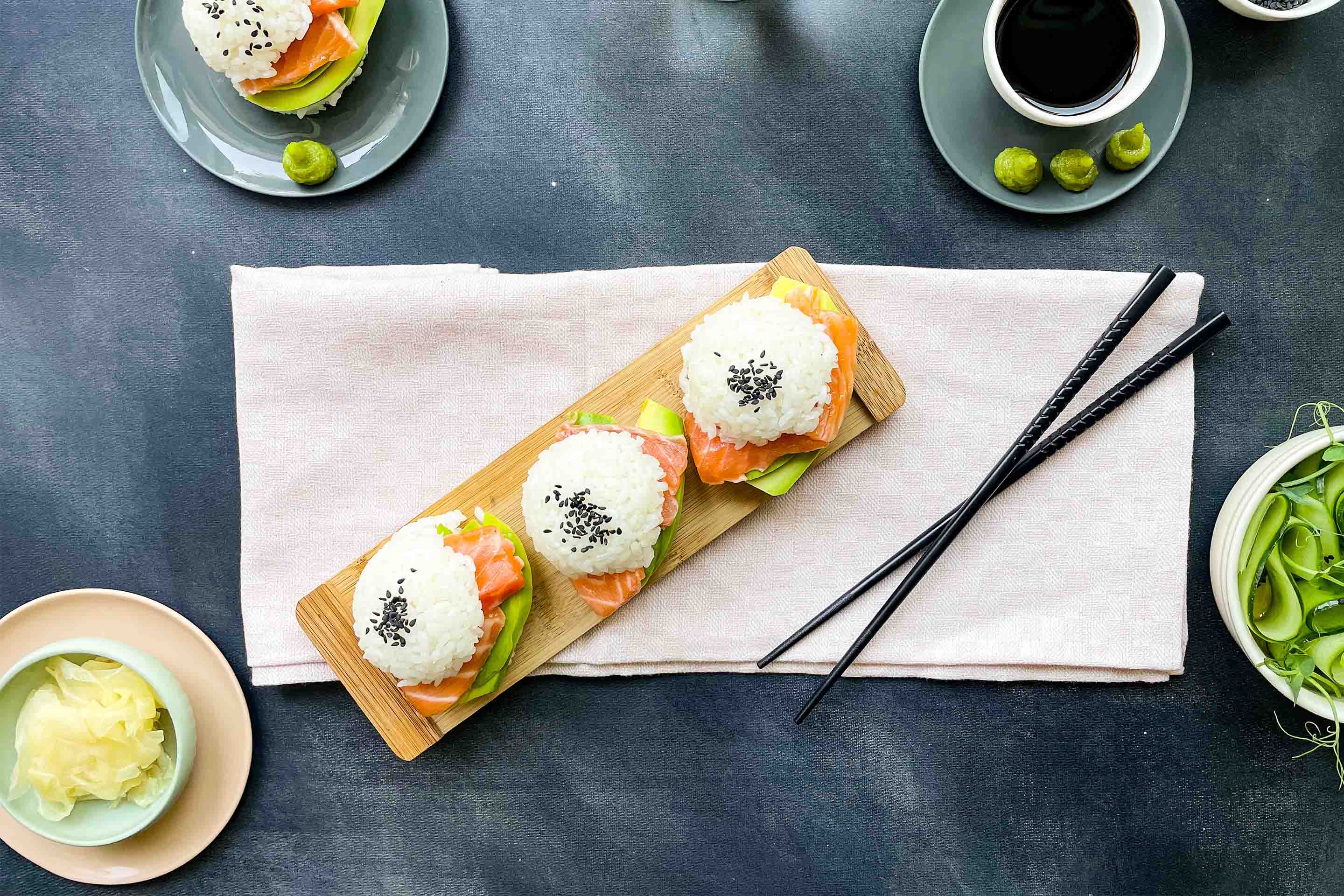 Три броя суши бургери върху дървена дъска и бяла кърпа, поднесени с клечки за ядене, соев сос и маринован джинджифил в купички.