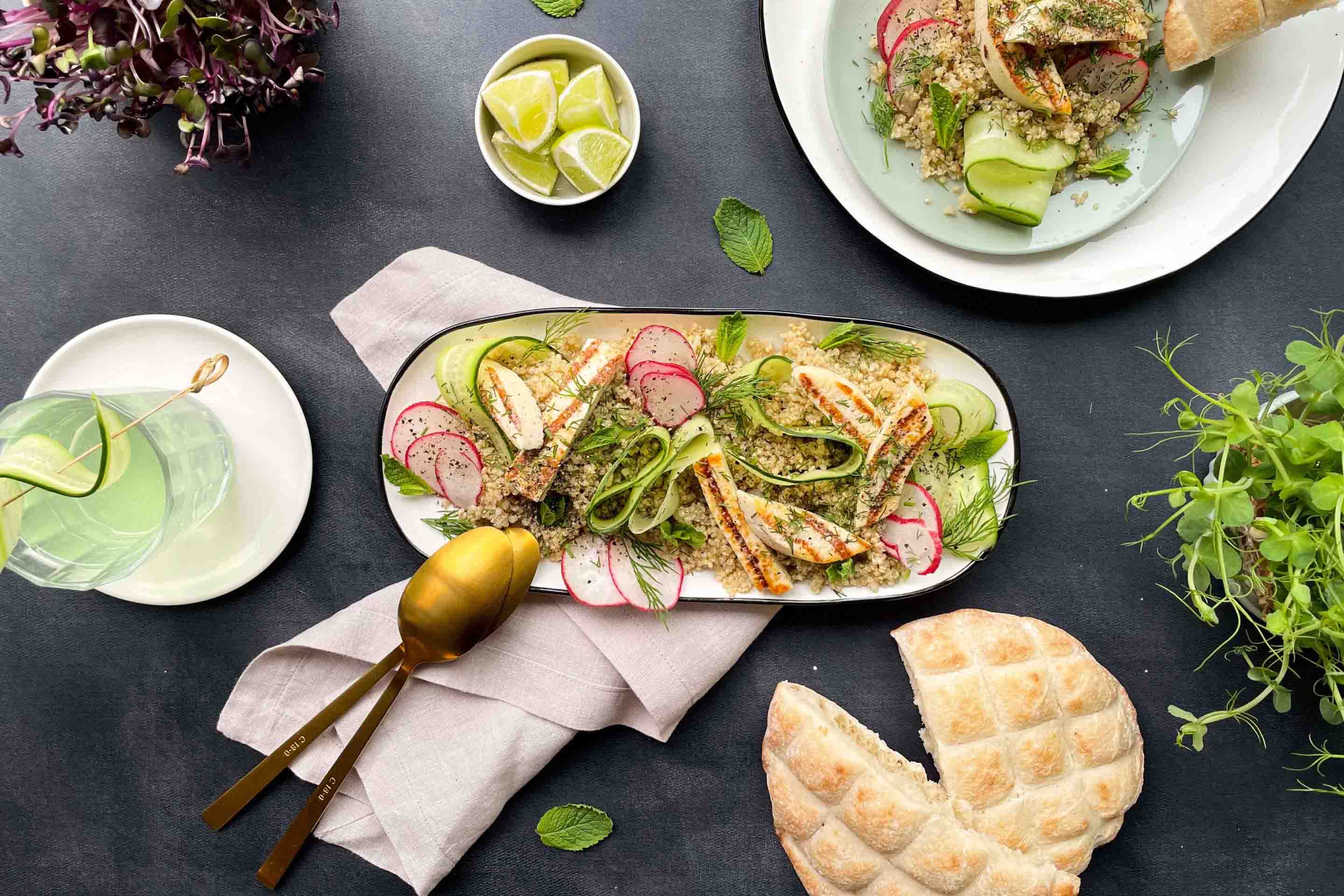 Салата в елипсовидна чиния с киноа, репички, краставици, грил сирене халуми и стръкчета копър, поднесена с лимонада и плоска питка.