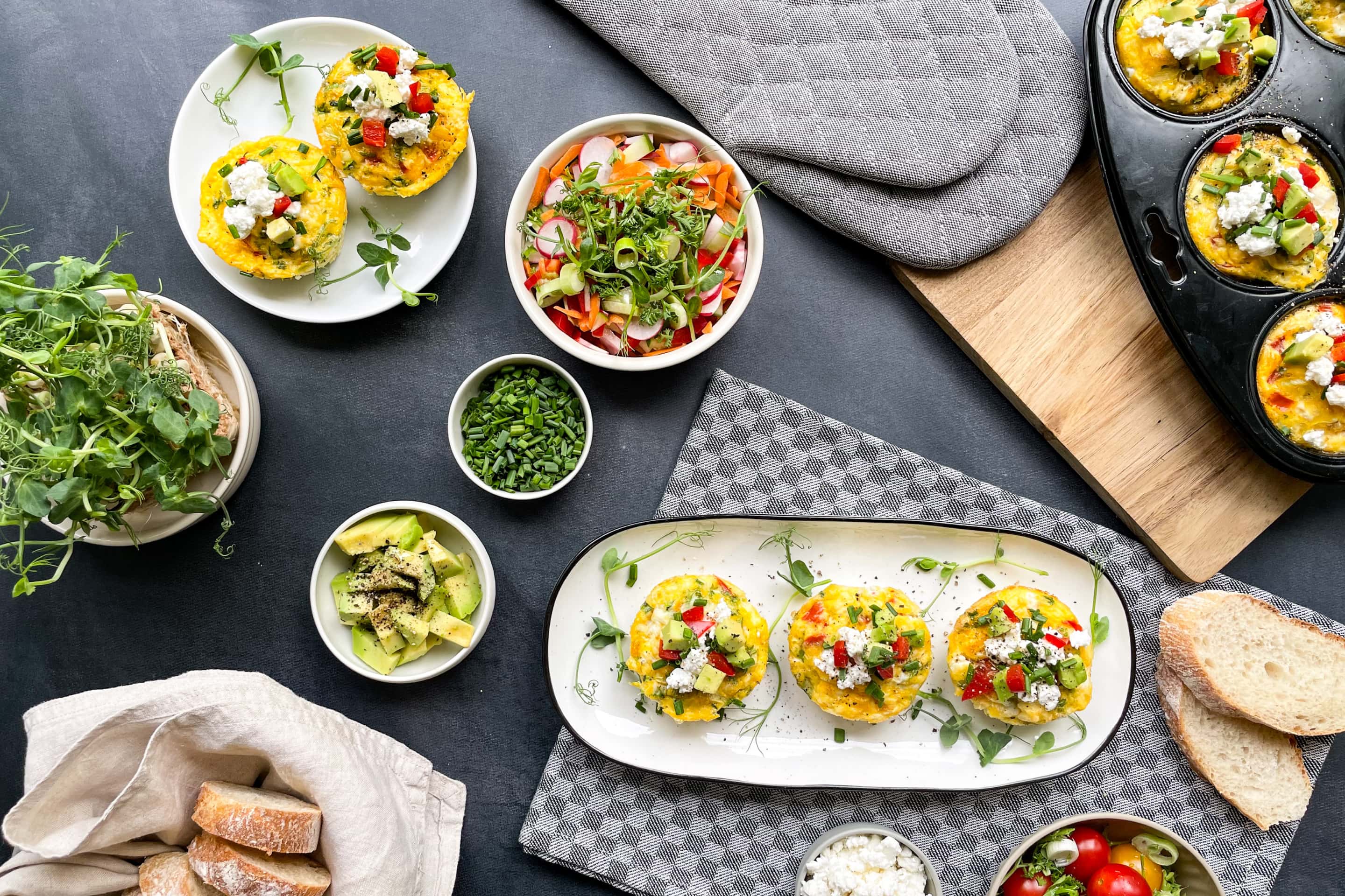 Солени мъфини от яйца със зеленчуци поднесени две в кръгла и три в елипсовидна чиния с добавки в разноразмерни купички от авокадо, див лук и морковена салата с репички и билки.