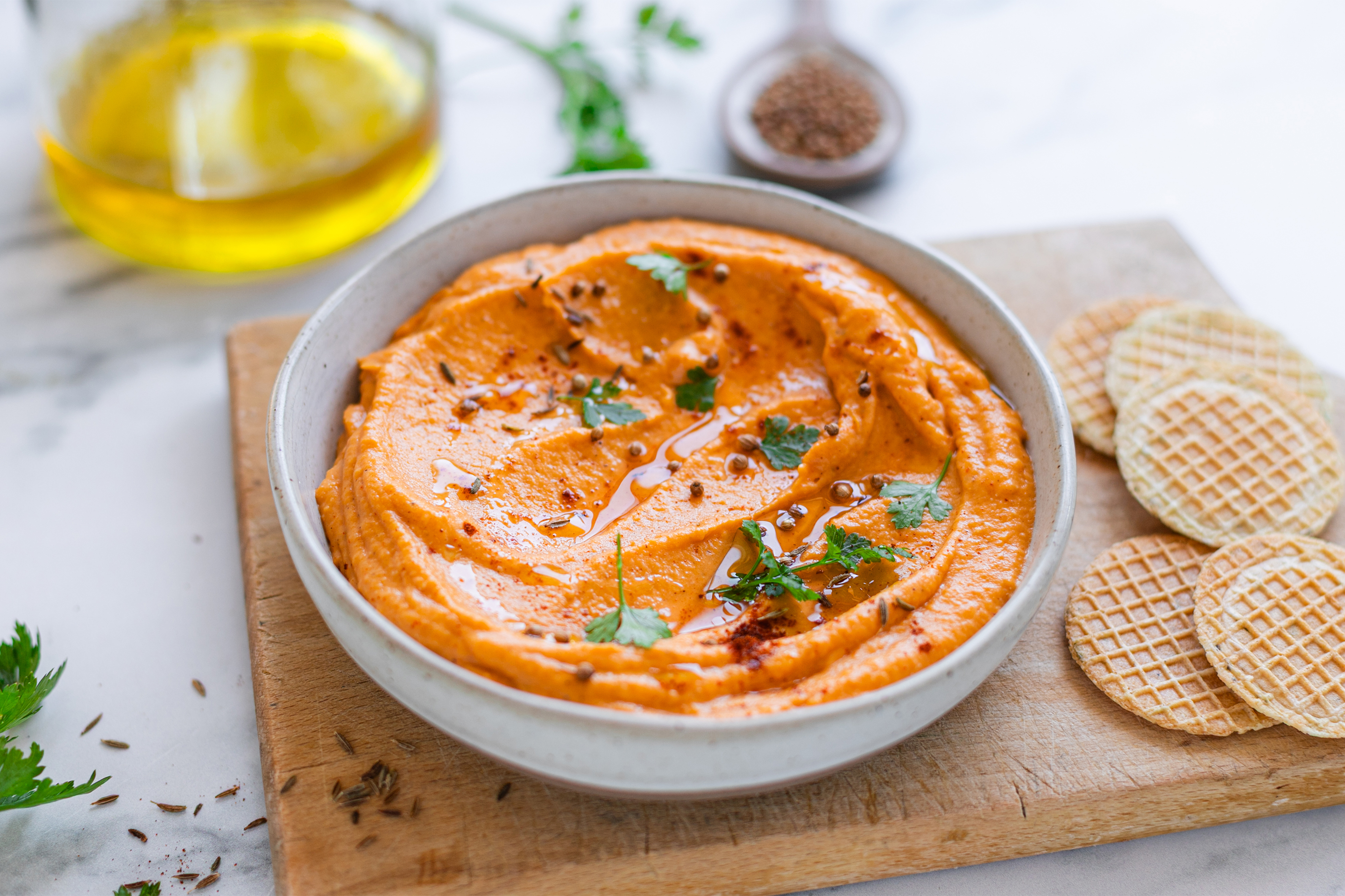 Оранжева разядка от сладки картофи, изсипана в кръгла и дълбока купа, с поръсен ситно нарязан магданоз, поднесена върху правоъгълна, дървена дъска с кръгли крекери.