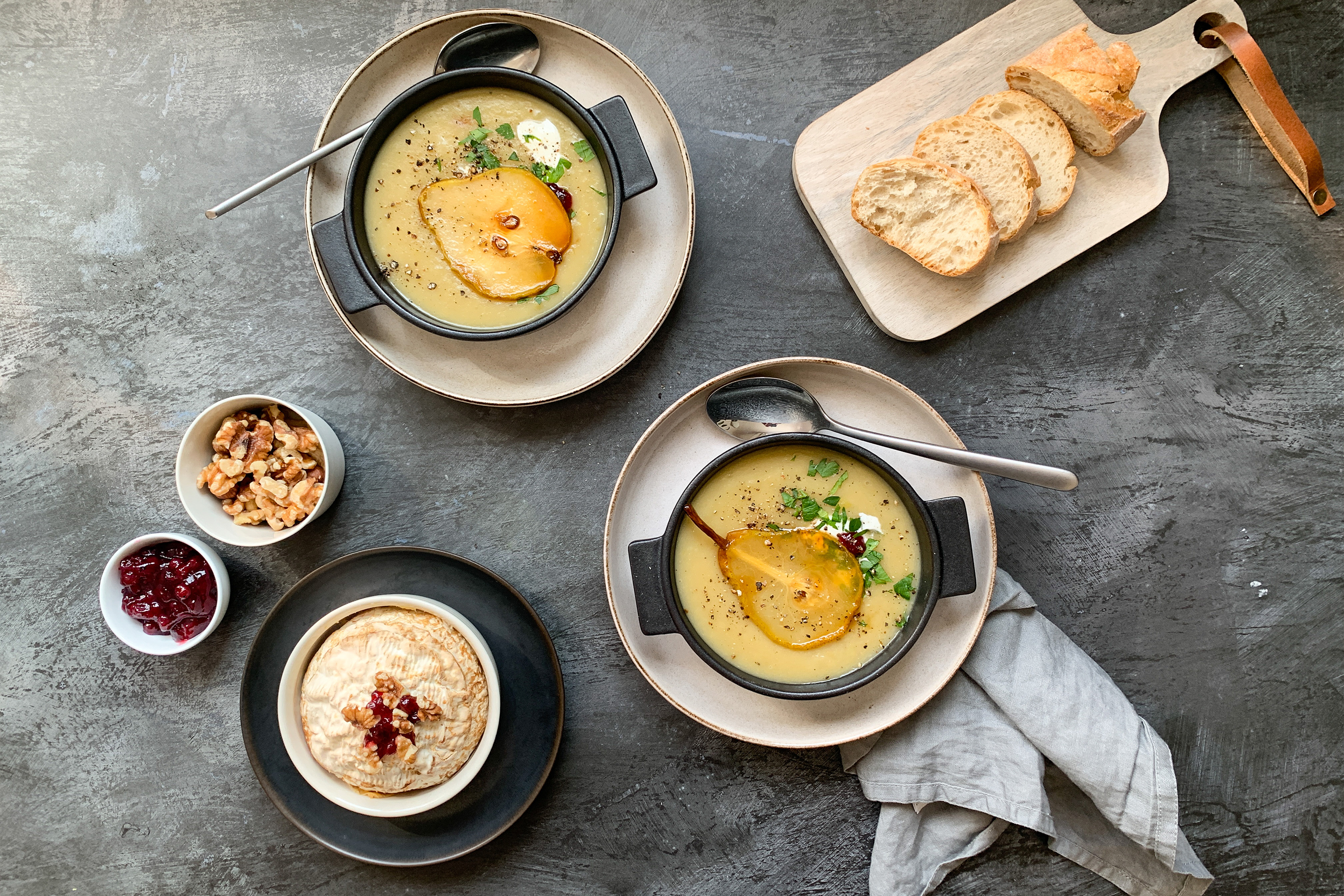 Две порции картофена крем супа с по резенче карамелизирана круша, поднесени с филийки хляб върху дървена дъска, орехи, стафиди и купичка с разтопено сирене Бри.