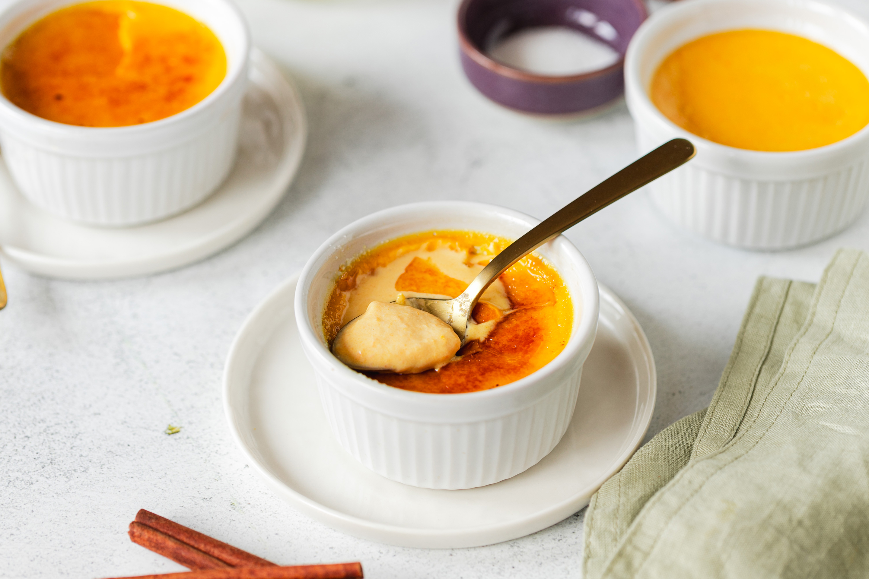 Три порции крем брюле, на преден план е порция с разчупена коричка и лъжичка.