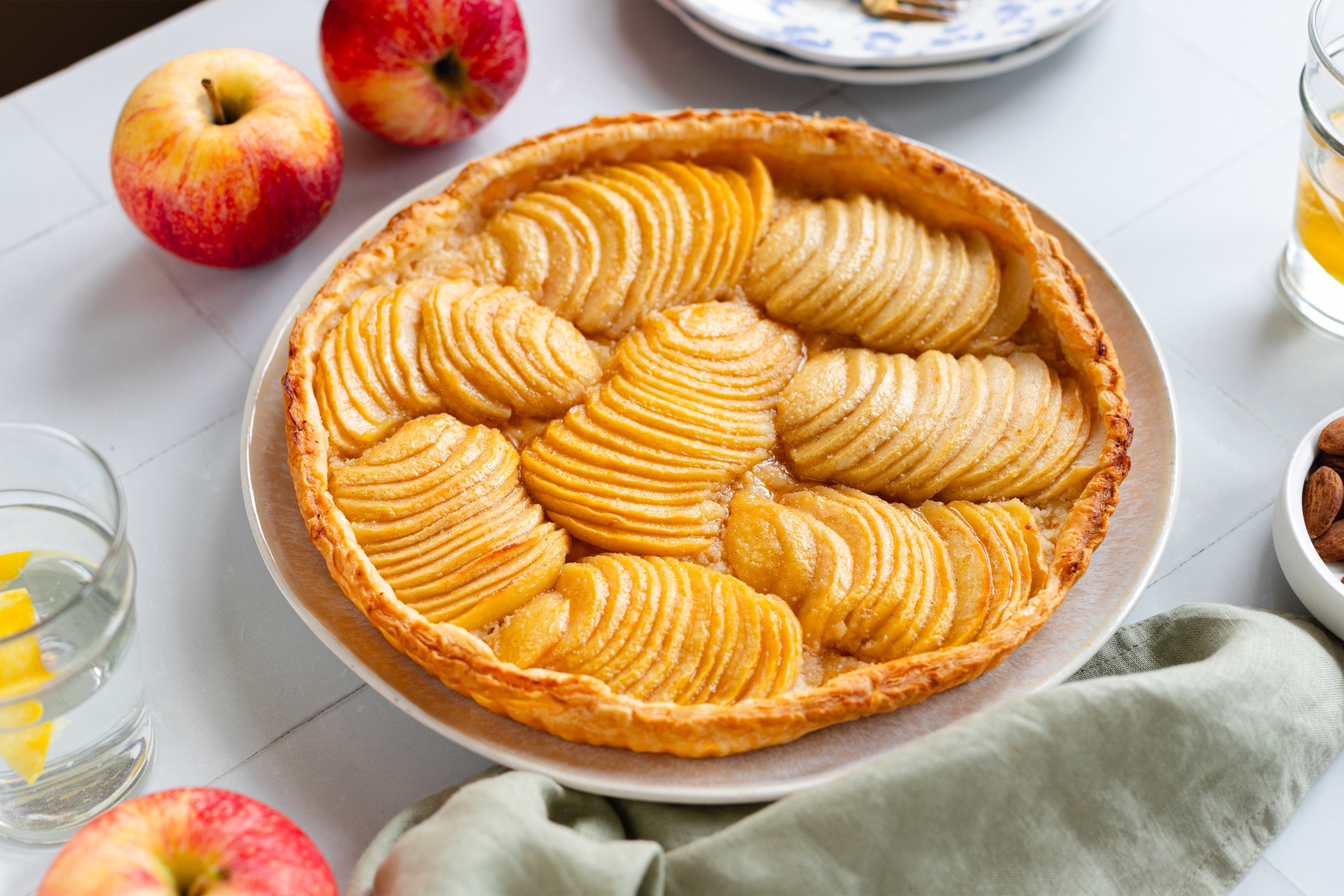 Голяма чиния с тарт с резенчета ябълки, аранжиран с пресен ябълки и купичка бадеми.