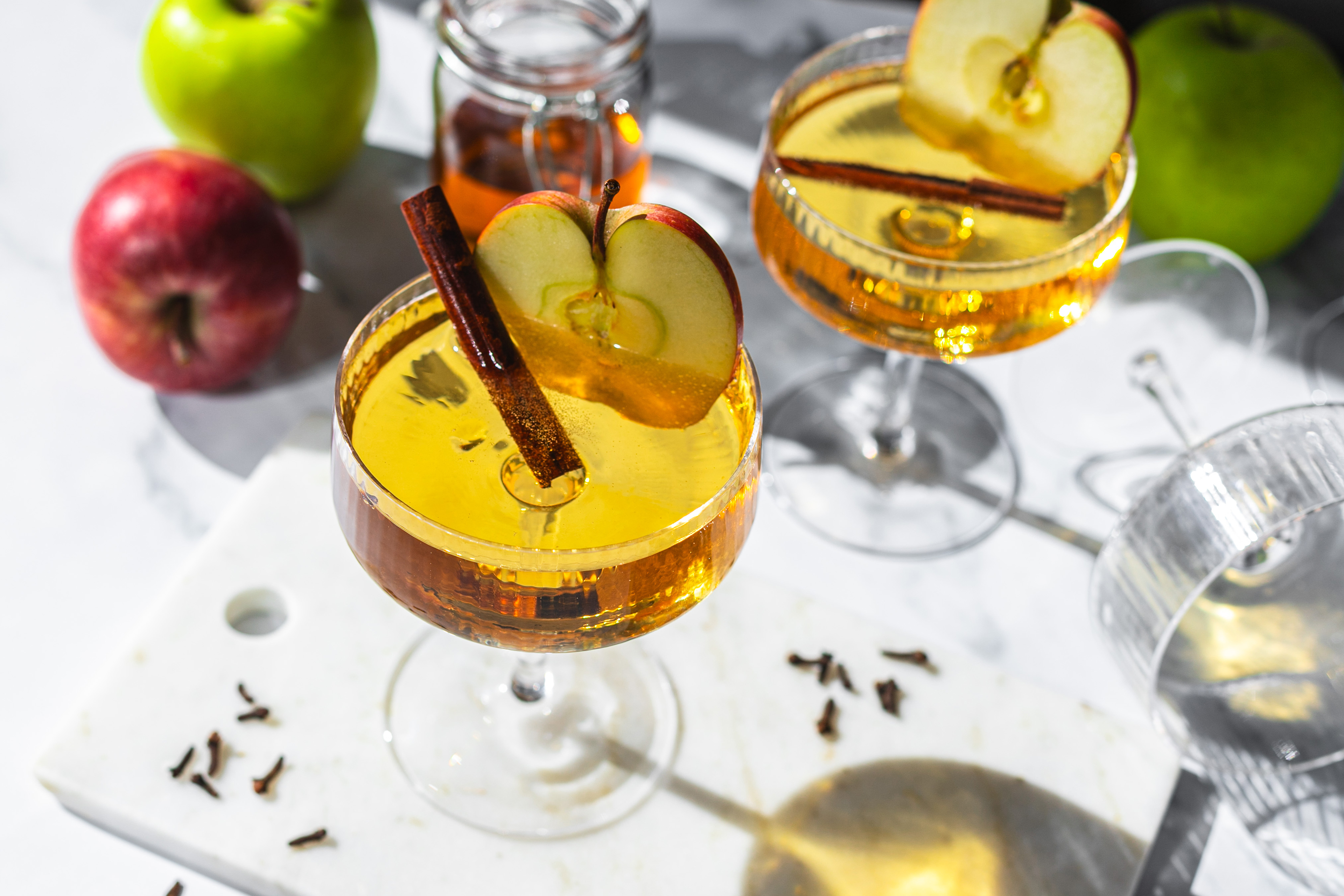 Два броя коктейли в купе чаши с по една канелена пръчица във всяка, аранжирани с ябълки и зрънца от карамфил.