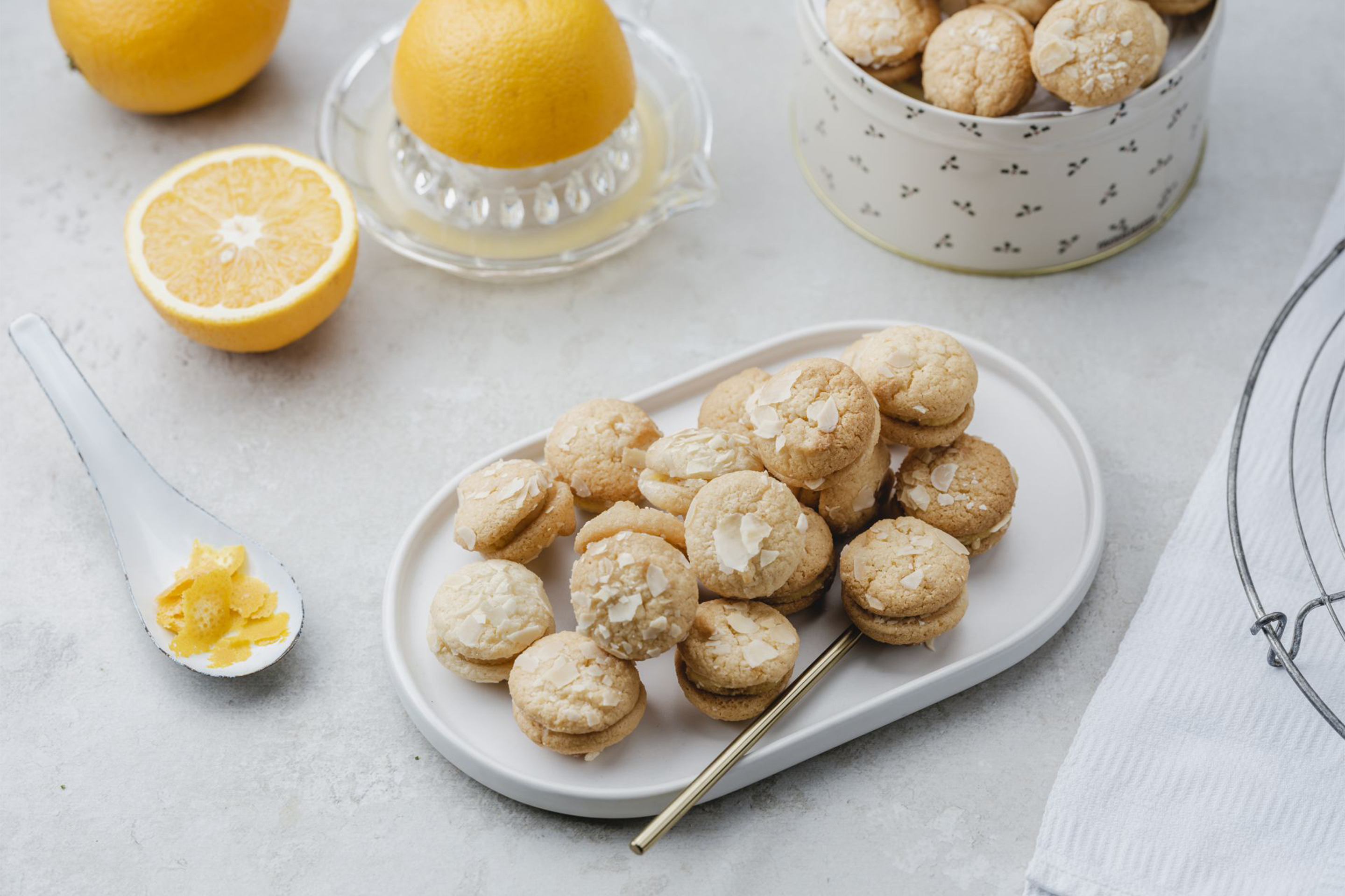 Порция, слепени две по две, светли курабийки с парченца филирани бадеми, поднесени в елипсовидна чиния в аранжировка с портокали на заден план.