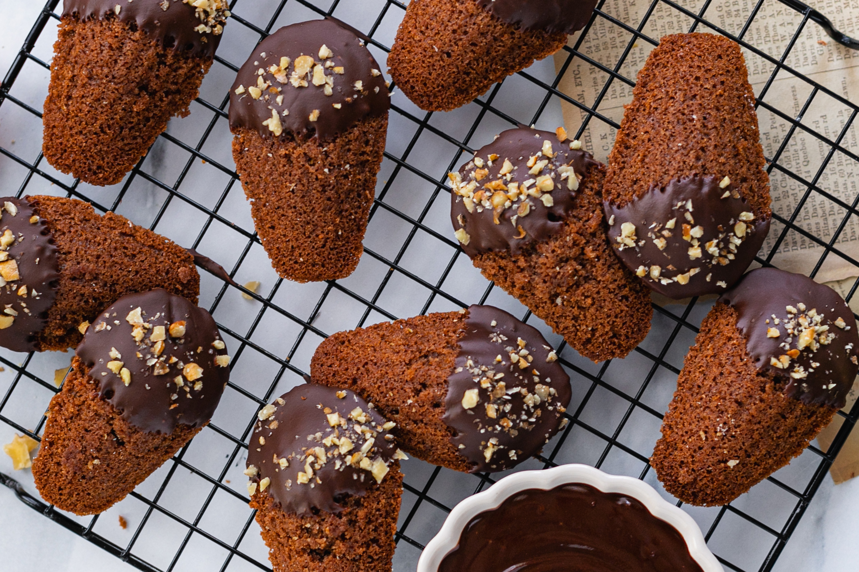 Осем броя шоколадови мадлени, чиито предни крайчета са били потопени в разтопен шоколад и поръсени с орехи, поднесени върху черна решетка и с купичка разтопен шоколад.